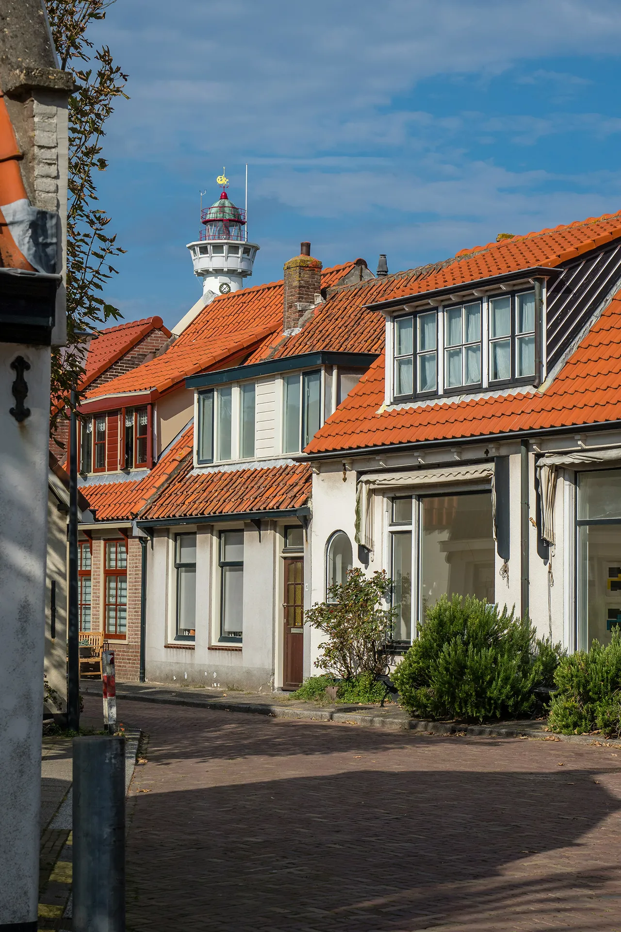 Straße mit Häusern und roten Ziegeldächern vor blauem Himmel und einem weißen Leuchtturm im Hintergrund