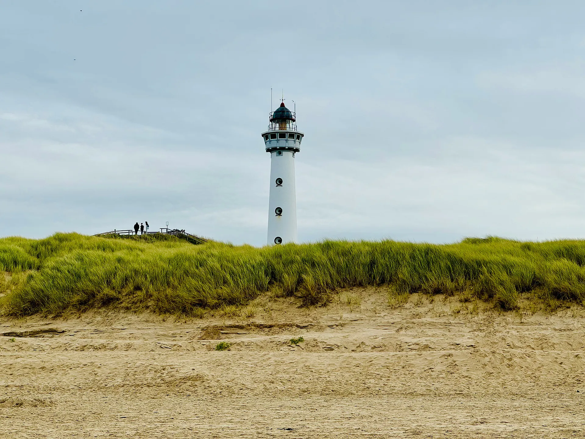 Leuchtturm hinter einer Dünenlandschaft mit Gras und Sand unter bewölktem Himmel