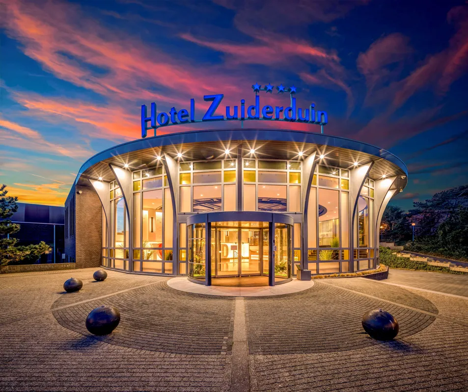 Aussenansicht eines runden Hotelfoyers mit großer Glasfassade und beleuchtetem Schriftzug "Hotel Zuiderduin" bei Sonnenuntergang.
