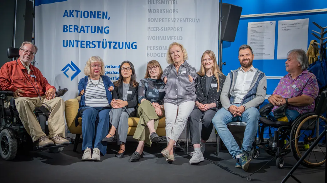 Gruppe von sieben Personen unterschiedlichen Alters sitzt in einem Raum mit blauem Hintergrund und Banner mit den Worten 'AKTIONEN, BERATUNG, UNTERSTÜTZUNG'