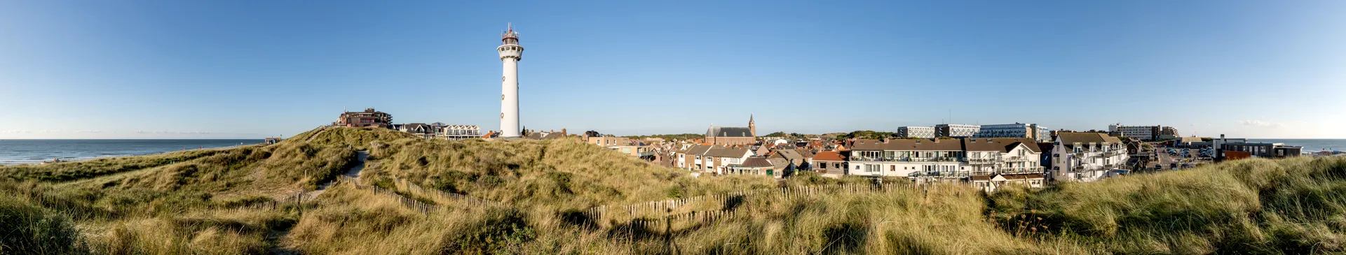 Leuchtturm auf einer Dünenlandschaft mit Häusern und einer Kirche im Hintergrund unter klarem Himmel