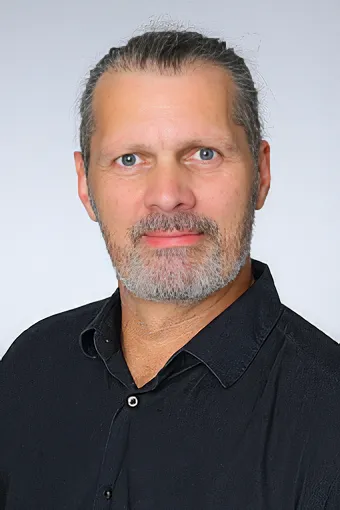 Niecke_Alexander A portrait photo of a man with his hair tied back and a full gray beard, wearing a black shirt.